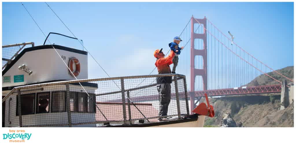 A man holding a toddler on a boat deck with the Golden Gate Bridge in the background under a clear blue sky.