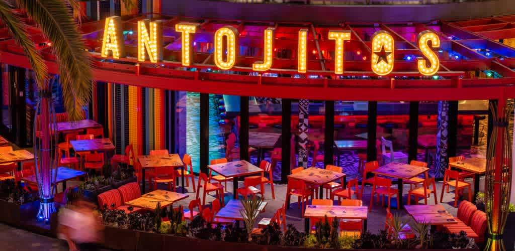 Colorful outdoor dining area with bright illuminated signage reading Antojitos, featuring tables, chairs, and vibrant decor in a lively atmosphere.