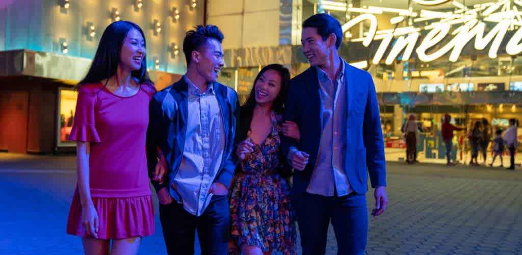 A group of four young adults is walking and laughing outside a theater at night with bright lights and a large illuminated sign in the background.