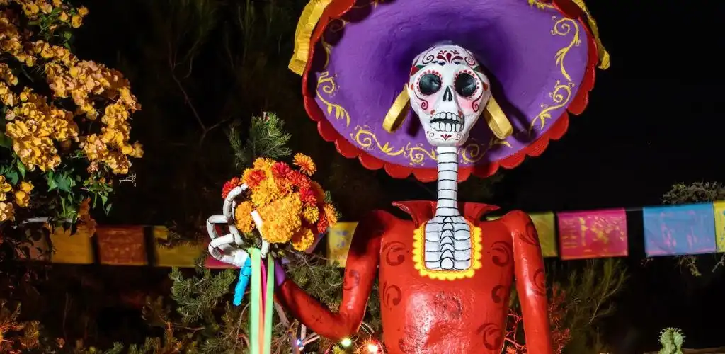 A colorful skeleton figure dressed in a red outfit with a large purple and yellow hat holds a bouquet of flowers against a dark background.