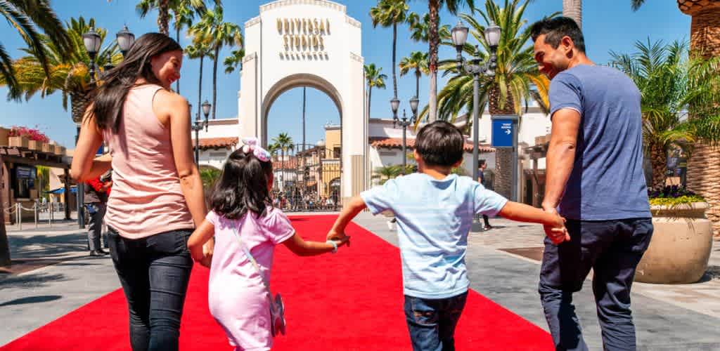 A diverse family walks hand in hand on a red carpet outside Universal Studios theme park under a sunny sky with palm trees.