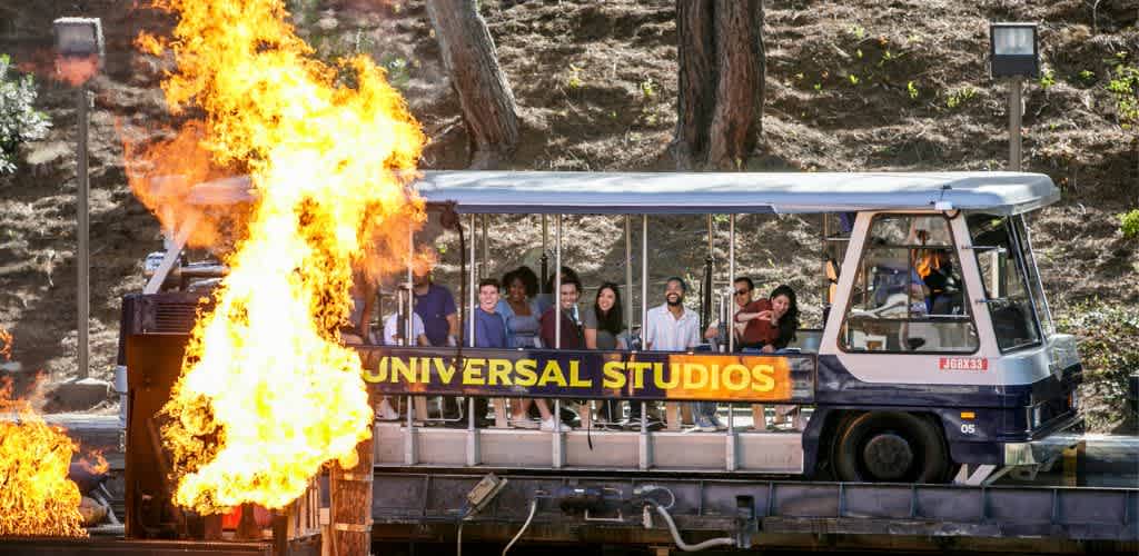 People riding a tram with a Universal Studios sign, surrounded by flames and explosion effects.
