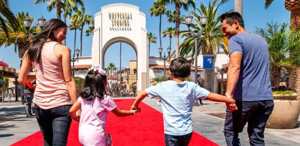 A family of four walking on a red carpet in front of a theme park entrance surrounded by palm trees and bright blue sky.