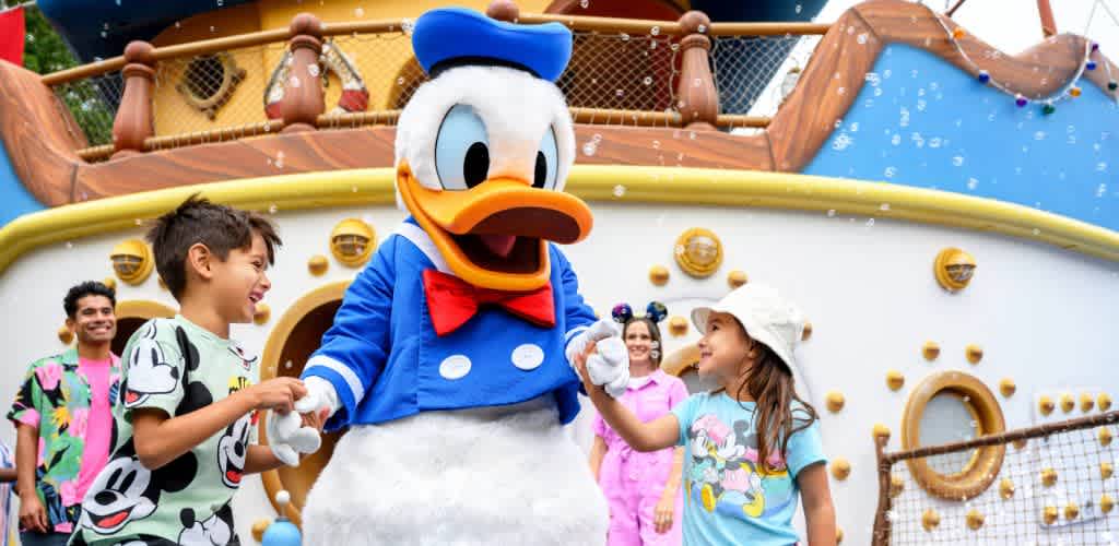 Children smiling and holding hands with a person in a Donald Duck costume outdoors at a colorful amusement park