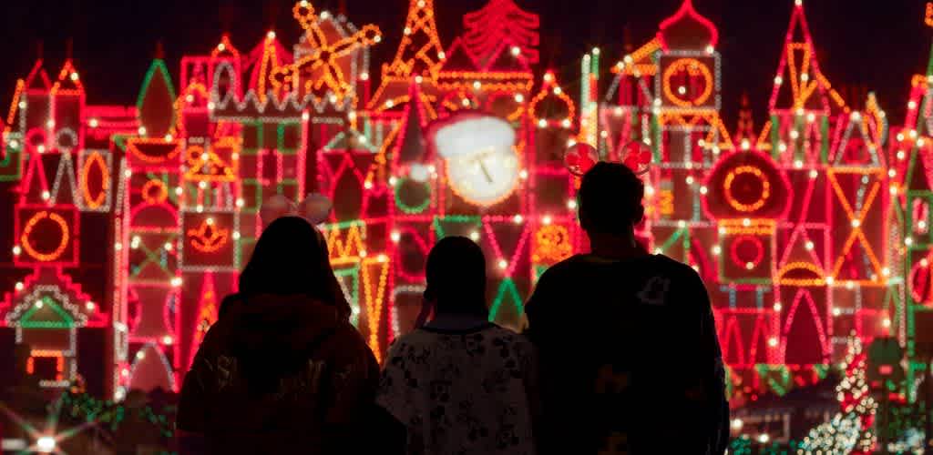 Three people look at colorful holiday lights display with illuminated buildings and festive decorations in the background.