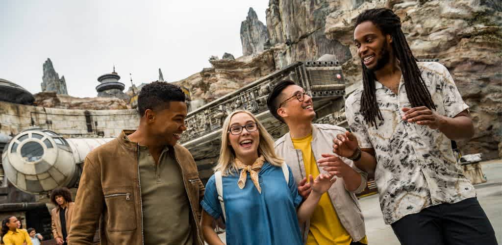 Group of diverse young adults smiling and talking outdoors at a themed park with futuristic aircraft and rocky landscape in the background