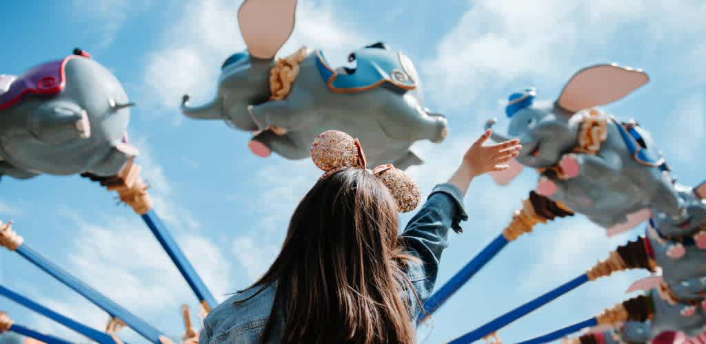 A girl with a bow headband reaching out towards a colorful carousel ride with elephant figures against a bright blue sky