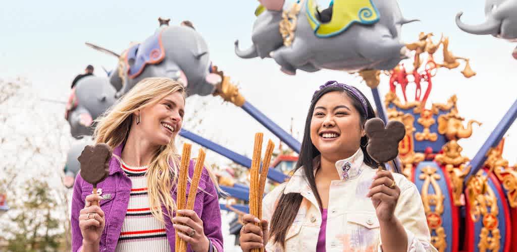 Two young women smiling and enjoying Churros and Mickey Mouse-shaped treats at a colorful amusement park with a carousel in the background