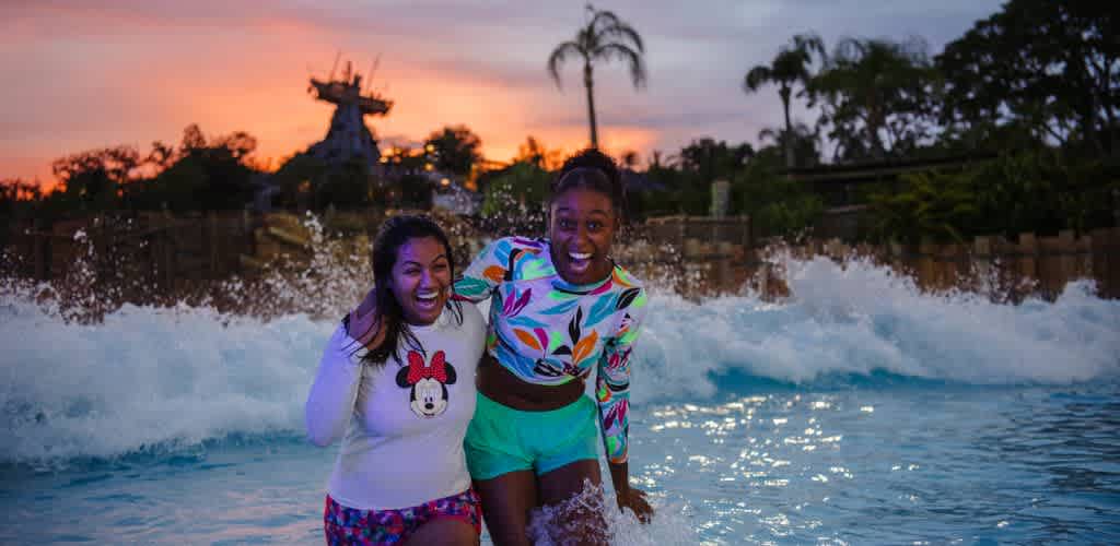 Two women smiling and enjoying themselves in a swimming pool at sunset with water splashing around them.