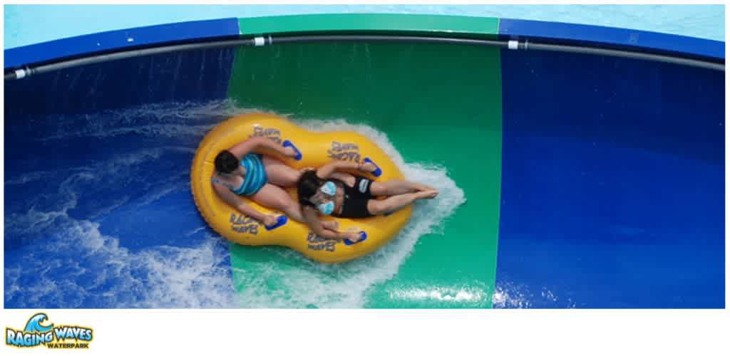 Two children wearing face masks ride a yellow inflatable tube down a water slide at Raging Waves Waterpark.