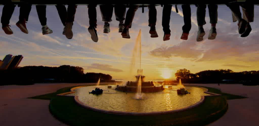 People's legs and feet are visible above a large fountain at sunset with trees and a colorful sky in the background