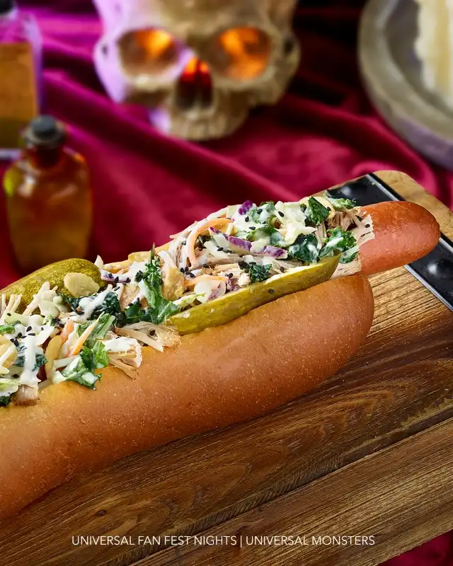 A hot dog with shredded chicken, chopped vegetables, pickles, and sauce in a bun on a wooden surface with a decorative skull in the background.
