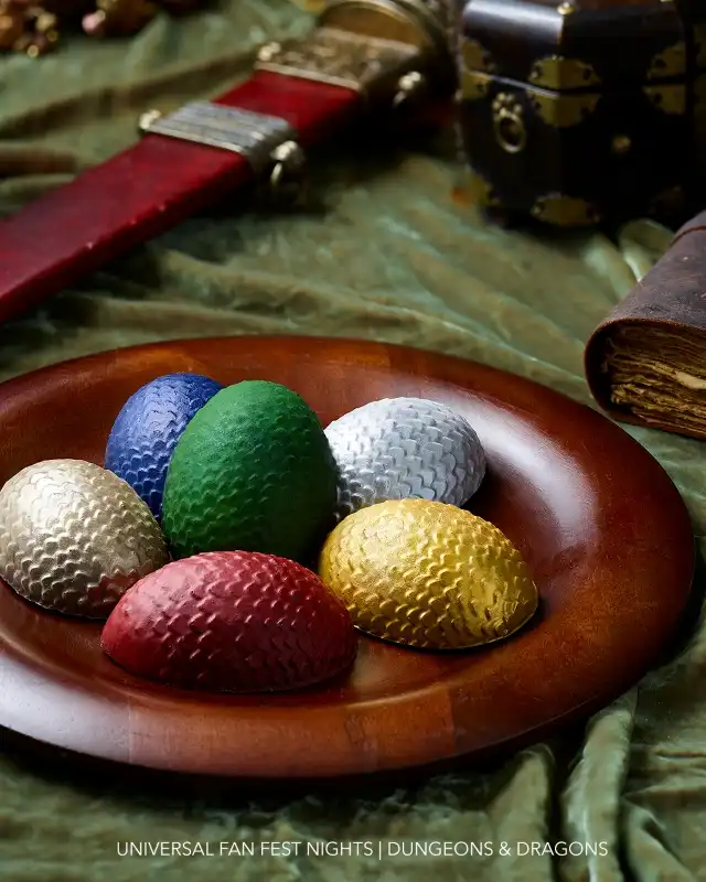 A wooden plate holds six colorful textured eggs including gold, silver, red, blue, green, and white, with gaming accessories and a green cloth in the background.