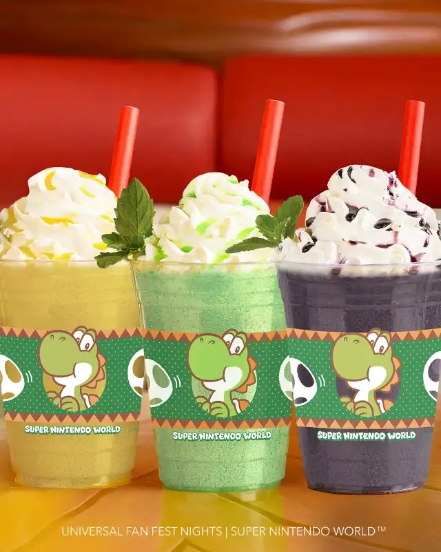 Three colorful drinks with whipped cream and mint leaves in Super Nintendo World cups on a yellow surface.