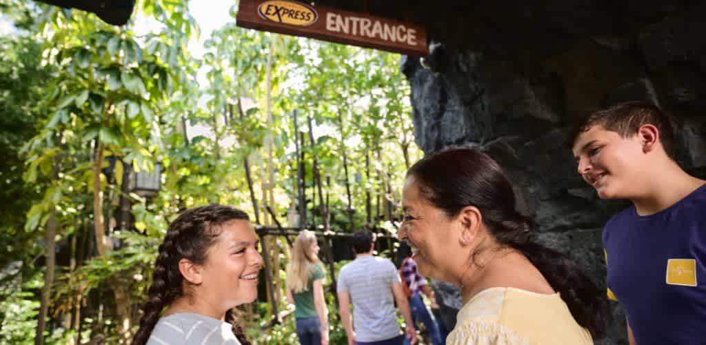 Two women and a young man smiling and talking outdoors near an entrance sign at a park or attraction.