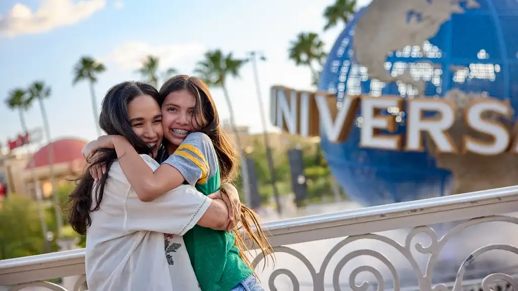 Two girls hugging happily in front of a Universal Studios globe sign during daytime outdoor visit.