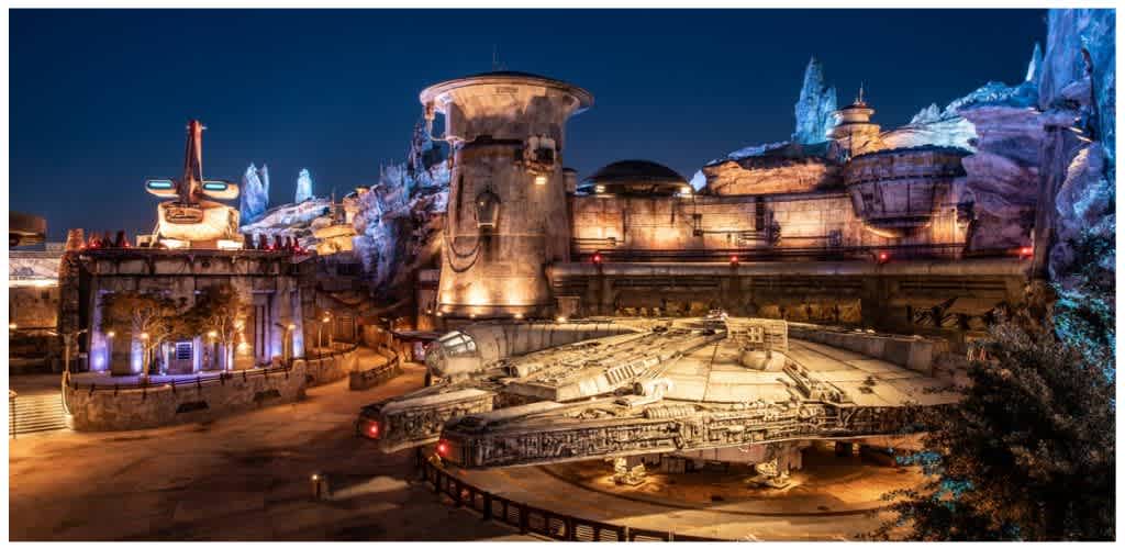 Nighttime view of a Star Wars themed area with a large Millennium Falcon spaceship replica and futuristic buildings illuminated with colorful lights.