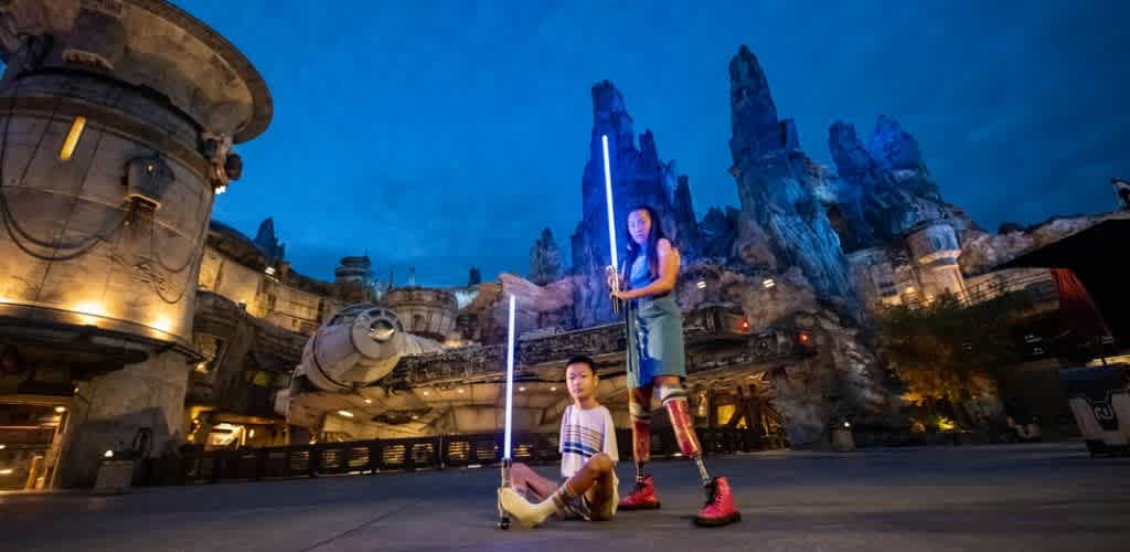 Two women with neon lights and laser weapons pose in front of a themed sci-fi building with rocky towers at night