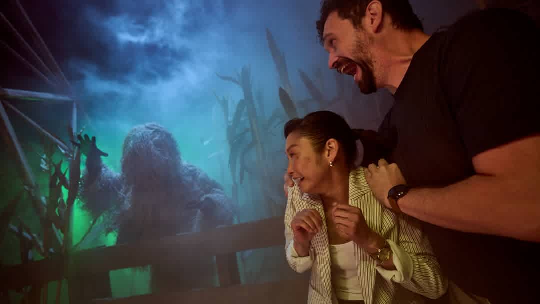 A woman and a man are excitedly watching a spooky swamp monster on a large screen. The scene is dark with eerie blue and green tones.