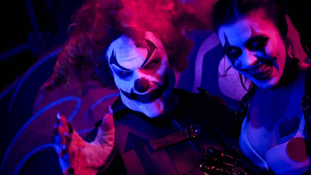 Two people dressed as scary clowns with painted faces and colorful costumes smile in a dark, neon-lit environment.