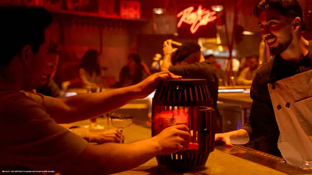 People sharing drinks at a lively bar with warm red lighting and a neon sign in the background.