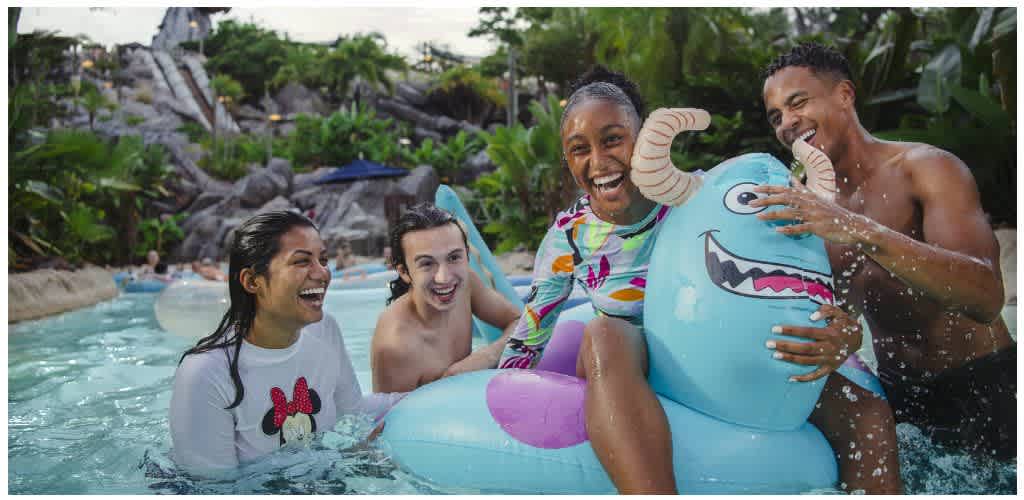 Four people enjoying a pool with a blue inflatable dragon float, smiling and having fun together in a lively outdoor setting.