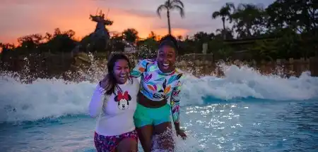 Two children enjoy playing and smiling in a water park with splashing waves and a giraffe sculpture in the background during sunset.