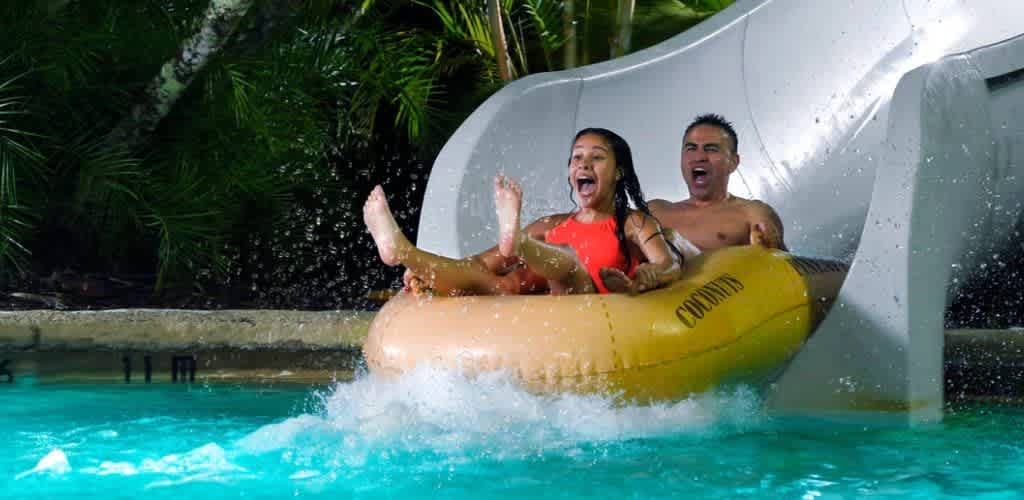 Children and adult enjoy riding down a water slide into a swimming pool with lush green plants in the background.