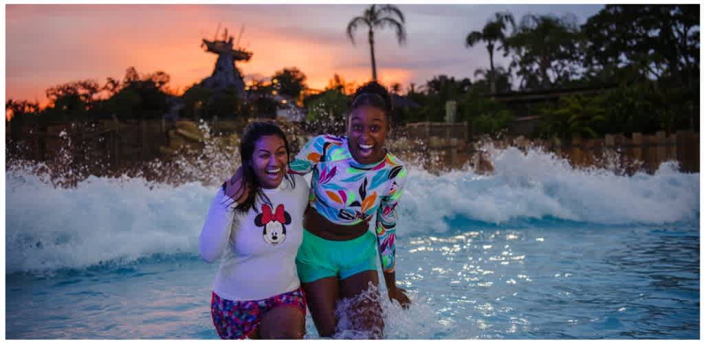 Two women enjoying themselves in a water park at sunset, smiling and splashing in the pool with a Disney-themed background.