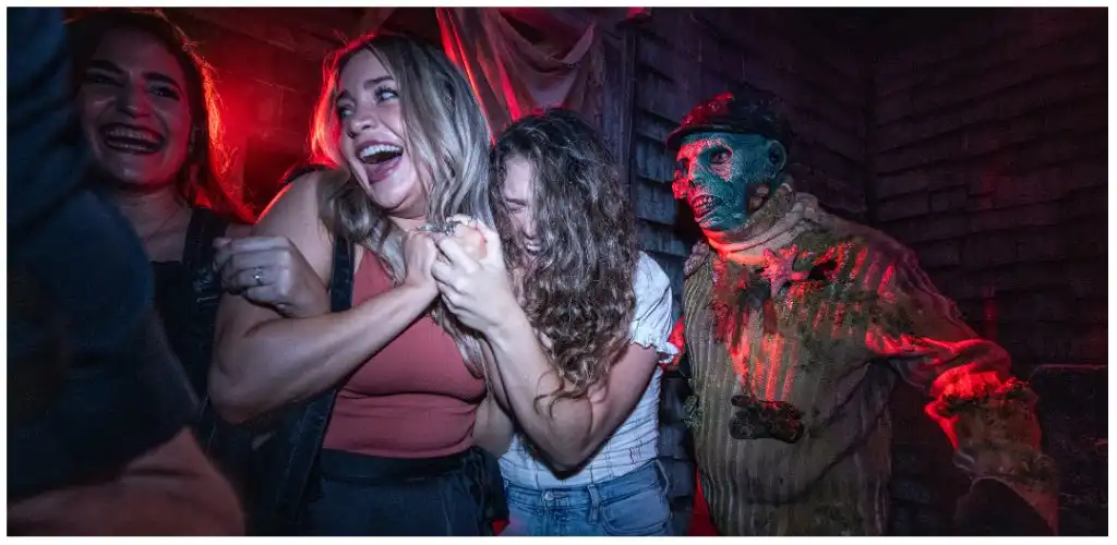 Group of smiling people having fun with a person in a zombie costume at a themed event in a dark, brick-walled setting
