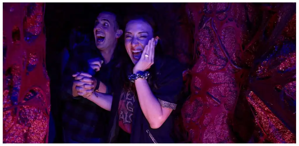 Two people are smiling and laughing inside a colorful, illuminated attraction with pink and purple lighting.