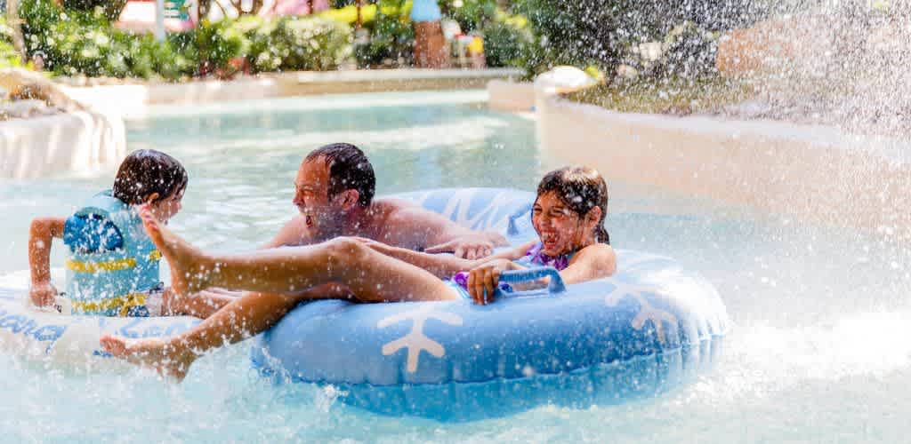 Children and an adult playing happily on a float in a pool on a sunny day with water splashing around