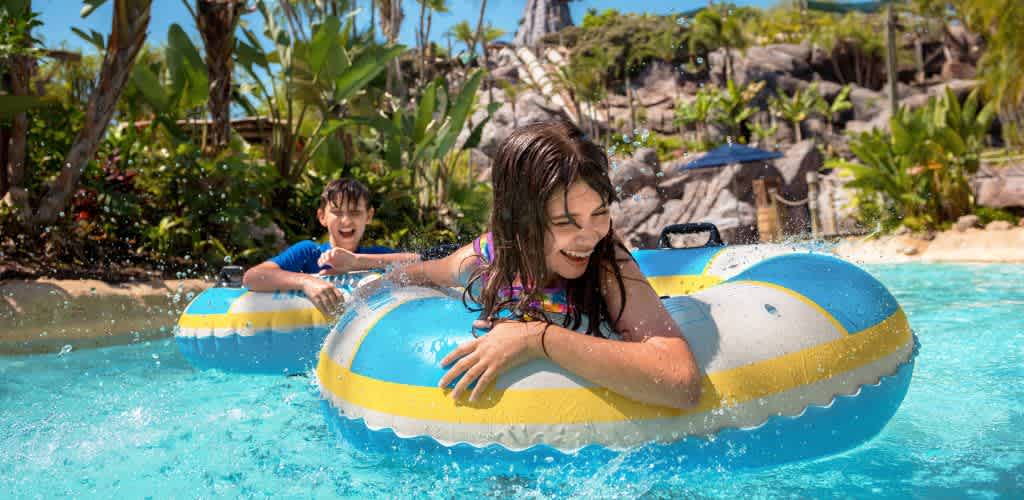 Two children happily play and laugh on a yellow and blue inflatable tube in a tropical pool surrounded by lush greenery and rocks.