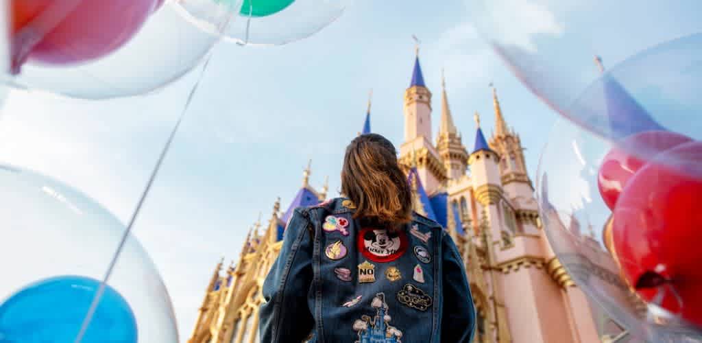 A person with shoulder-length hair wearing a jacket decorated with patches faces a Disneyland castle surrounded by colorful balloons in the sky.