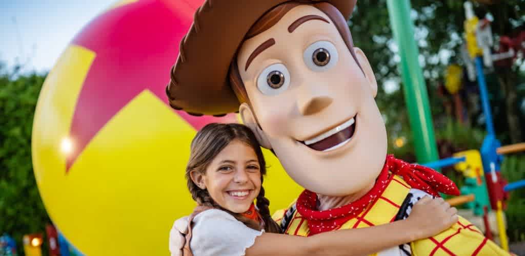 Child hugging animated Toy Story character in outdoor amusement park setting with colorful rides and a large yellow and red ball in the background