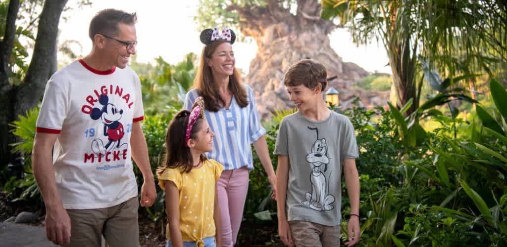 A family of four walking outdoors in a lush garden with a large tree in the background. They are smiling and enjoying each other's company.