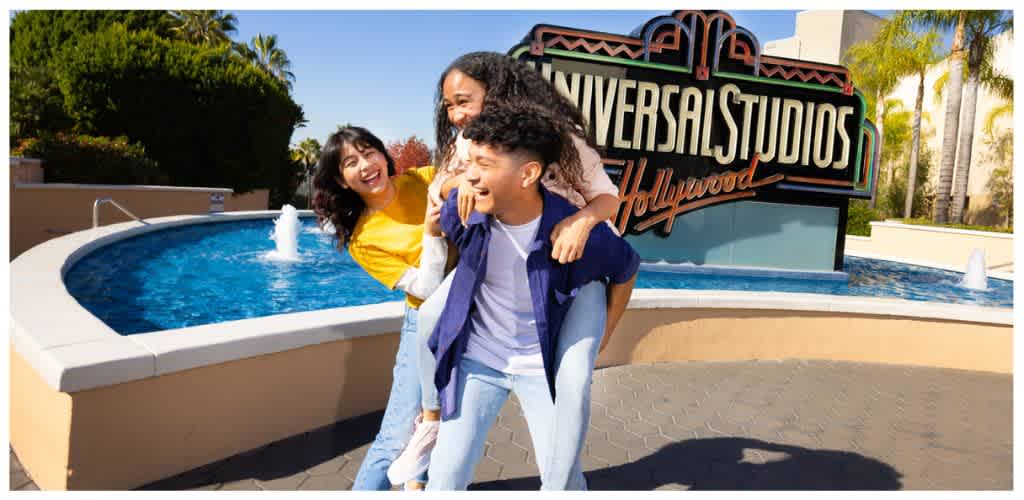 Three smiling young people standing in front of Universal Studios Hollywood sign outside a water feature with trees and a clear sky in background