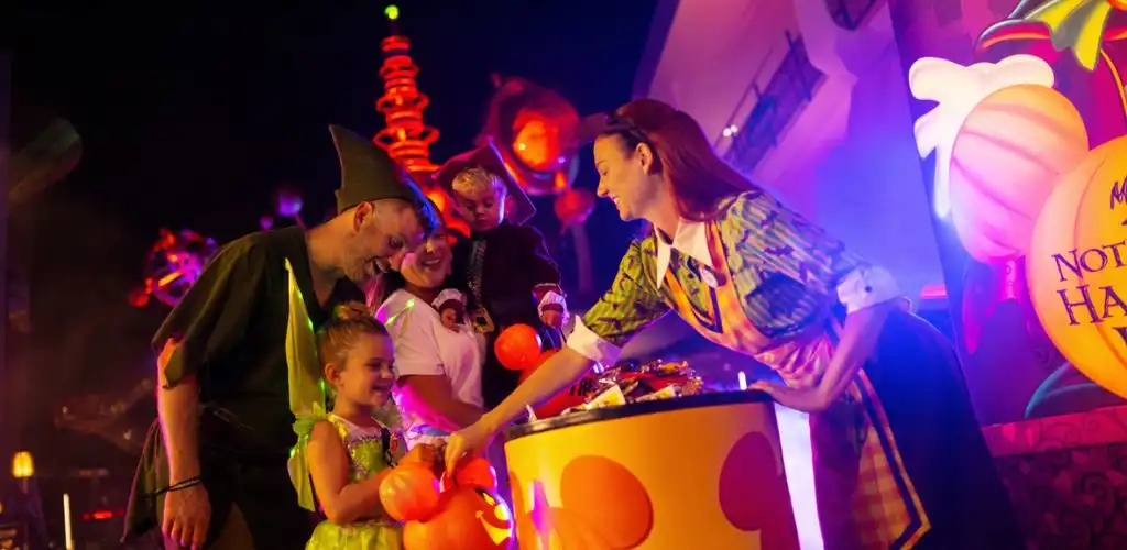 A group of children and adults in halloween costumes gather around a pumpkin-shaped container during a festive event