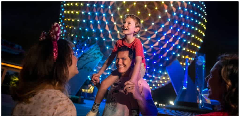 A smiling man carries a boy on his shoulders while a woman and another girl look on at a colorful amusement park with bright lights.