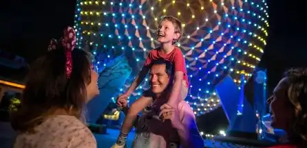 A joyful young boy sits on a man's shoulders while a woman smiles at them, all enjoying a nighttime amusement park with colorful illuminated rides in the background.