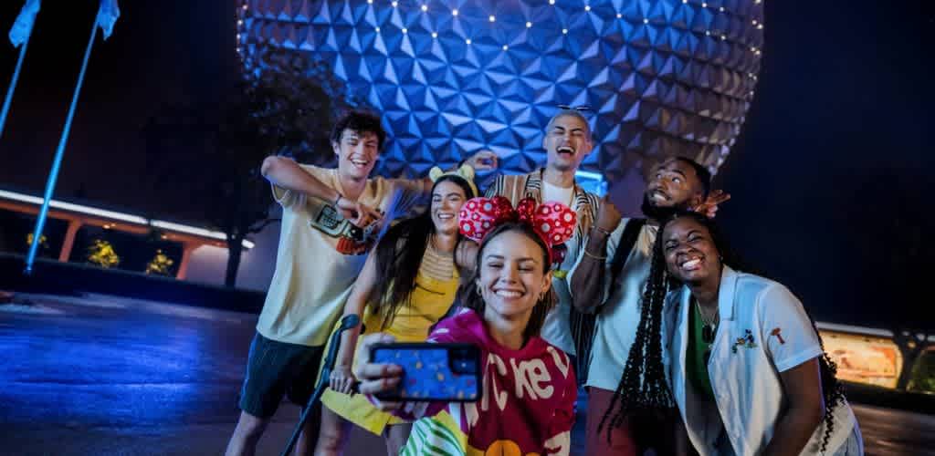 Group of diverse friends taking a selfie at night in front of spaceship earth at EPCOT center glow with excitement and smiles