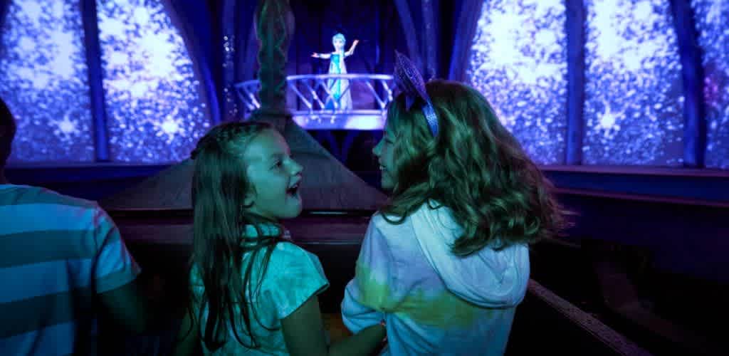 Two young girls laughing and talking while watching a performer on stage with snowflake patterns and blue lighting in the background.