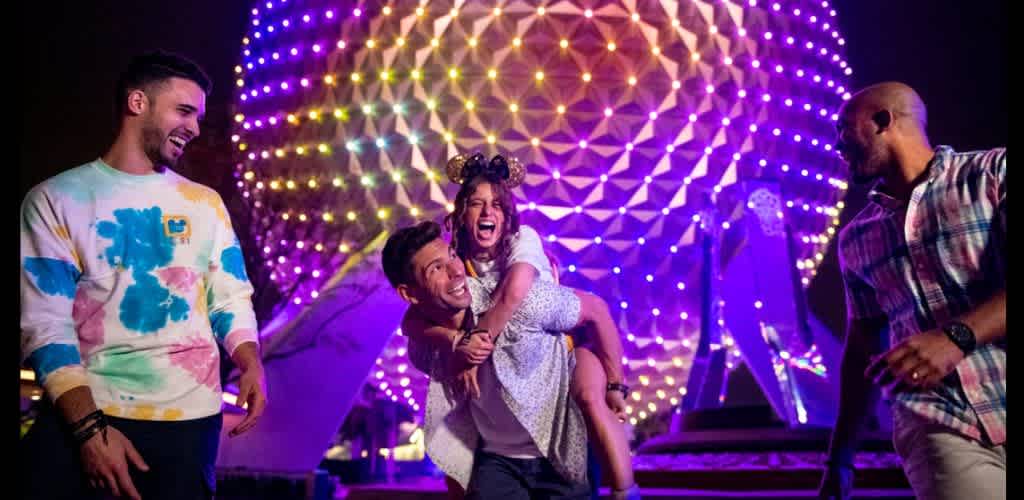 Group of four diverse people smiling and enjoying themselves at a colorful outdoor night event with bright lights and a large illuminated geometric structure.