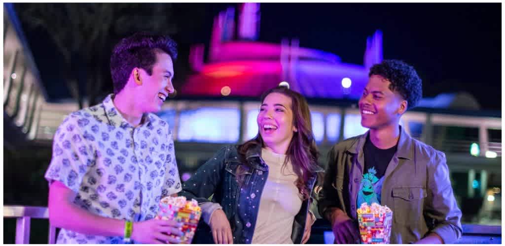 Three young adults smiling and chatting while holding popcorn bowls at a festive outdoor event at night