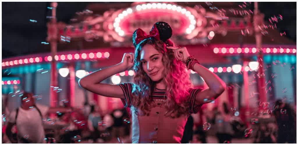 A young woman with curly hair wearing Minnie Mouse ears at an amusement park nighttime with colorful lights and a carousel in the background.