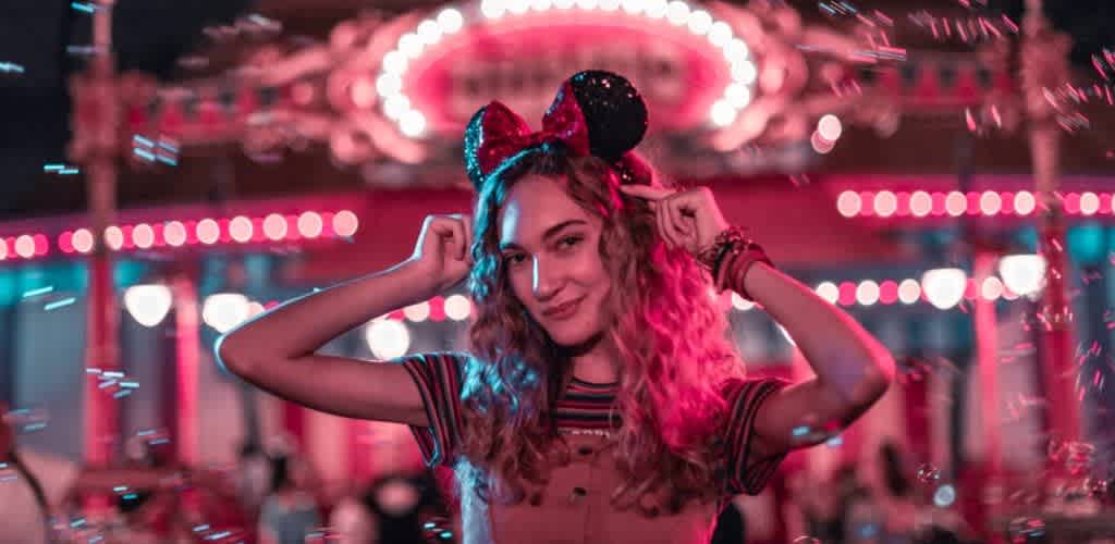 A young woman wearing Minnie Mouse ears at a colorful amusement park ride at night with bright lights in the background.