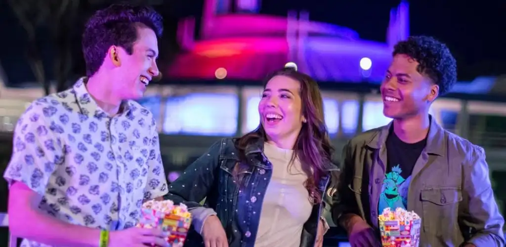 Three young adults smiling and holding popcorn at an outdoor event during evening with colorful lights in the background
