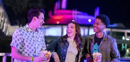 Three friends smiling and enjoying popcorn at night in front of colorful illuminated amusement park rides