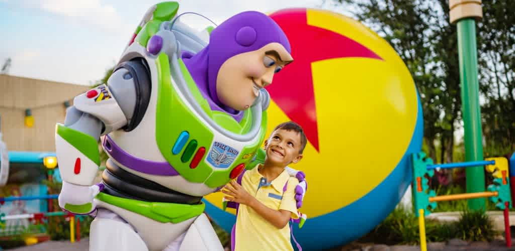 A young boy smiles and interacts with buzz lightyear at a colorful amusement park with Toy Story themed decorations and attractions.
