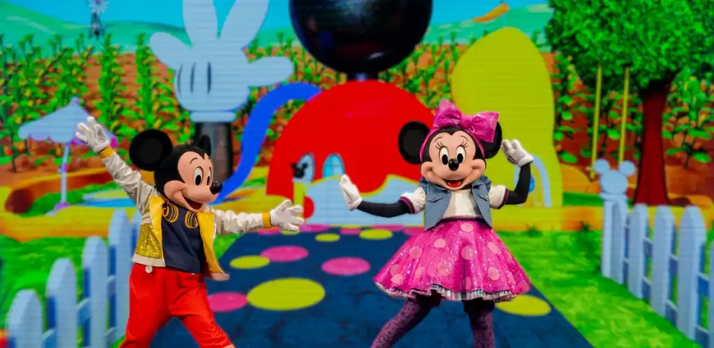 Mickey Mouse and Minnie Mouse characters standing outdoors on colorful pathway with green trees and whimsical background in a theme park setting